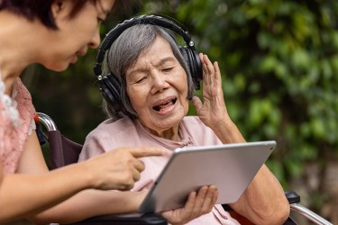 A person wearing headphones sings with their left hand up as another person points to a tablet
