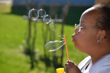 A person wearing glasses outside on a grassy area blowing bubbles